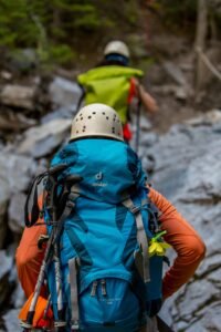 Two hikers with backpacks and helmets navigating a rocky trail in the forest.